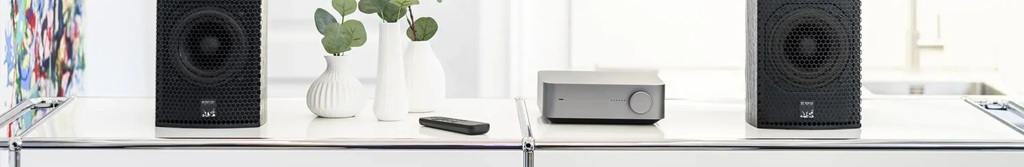 WiiM Amp Streaming-Verstärker zwischen zwei HiFi-Lautsprechern mit Fernbedienung im modernen Wohnraum.